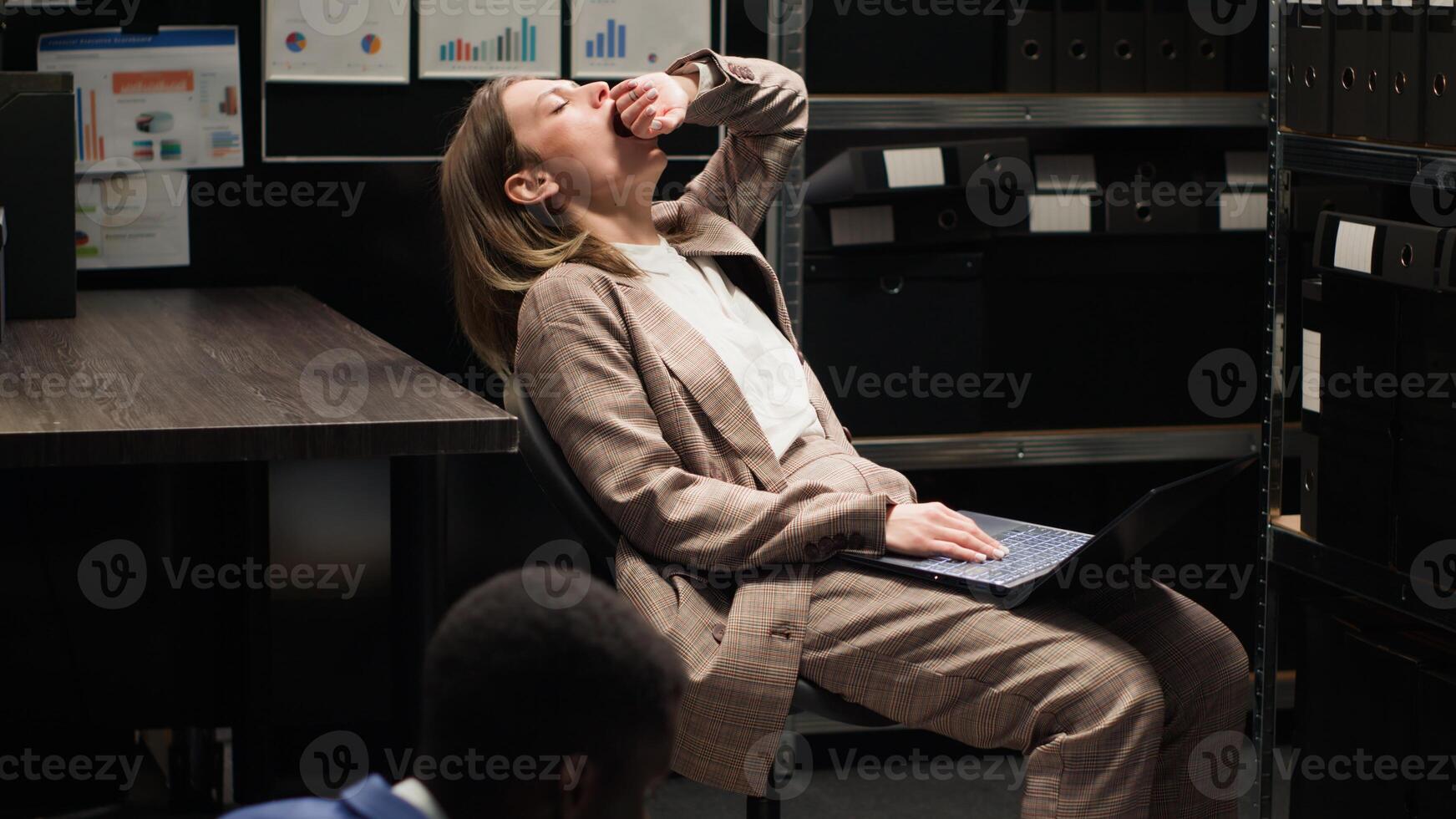 African american male officer and caucasian female inspector with laptop analyze evidence in incident room. Tired policewoman sits on chair while policeman on the floor examining clues and statements. photo
