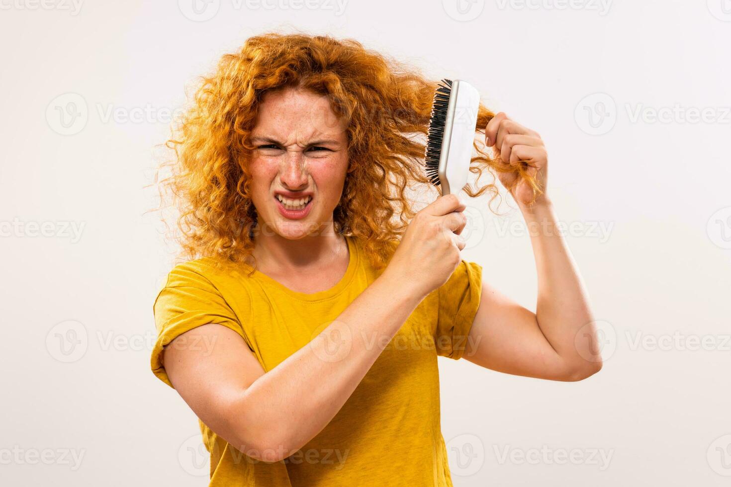 Angry ginger woman holding hairbrush and combing her curly hair photo