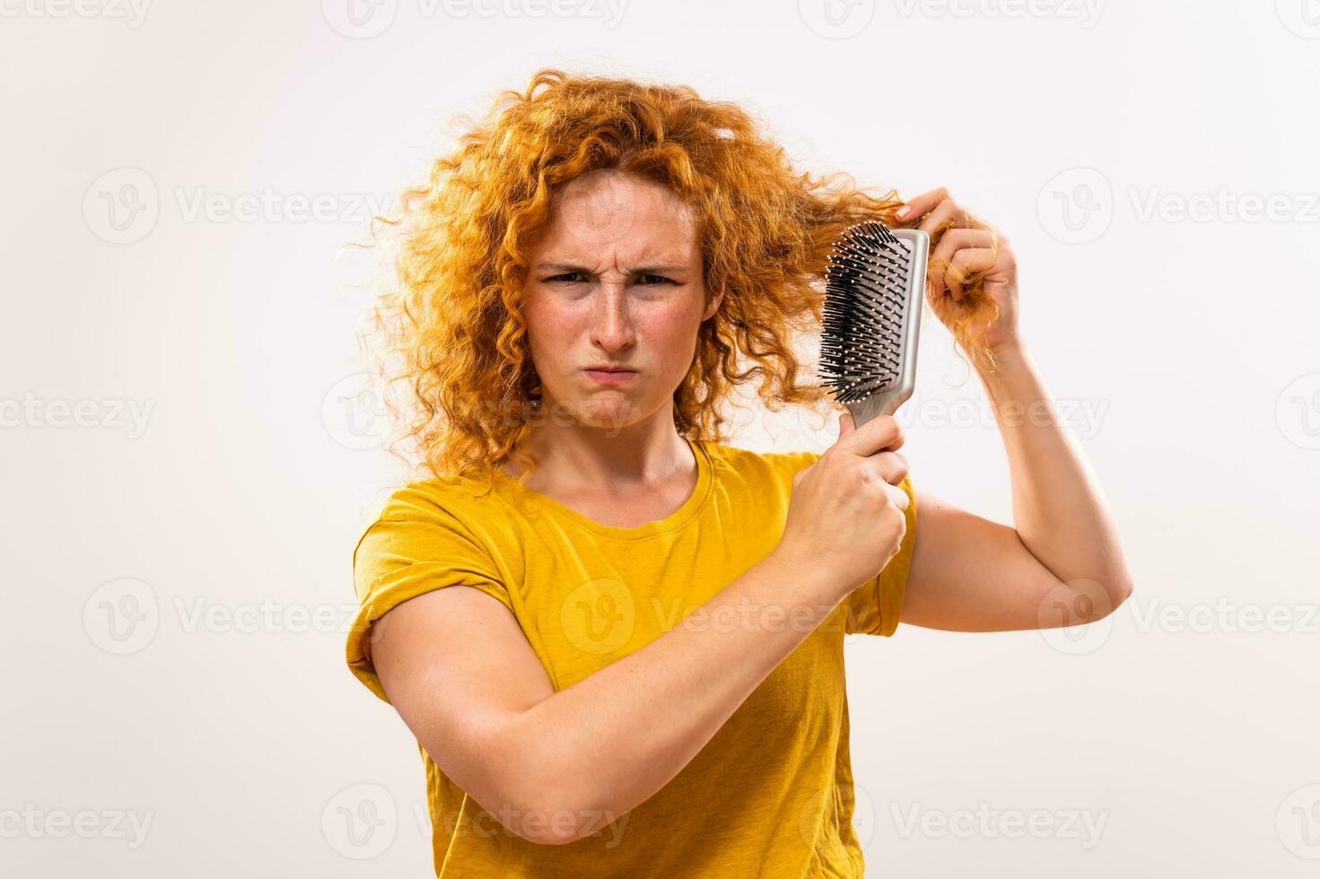 Angry ginger woman holding hairbrush and combing her curly hair photo