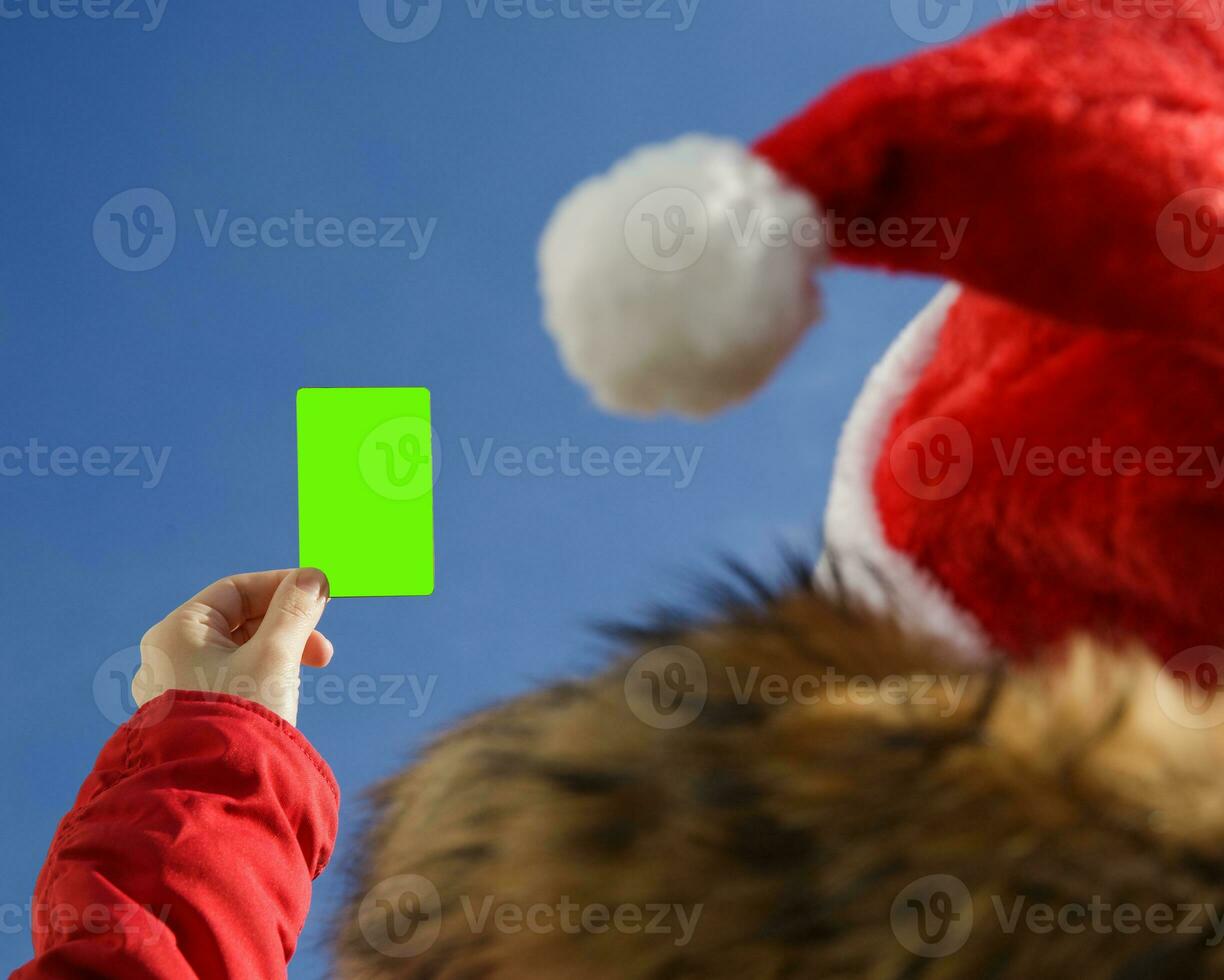 Closeup of a woman's hand holding a blank green gift card.in Santa hat and red jacket. gift or discount concept.High quality photo.selective focus photo