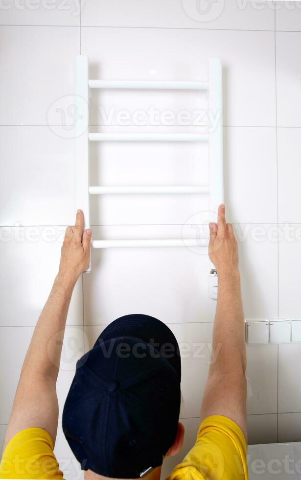 Renovation in the bathroom. A plumber in a cap and yellow shirt installs a heated towel rail. Interior walls with light tiles. High quality photo