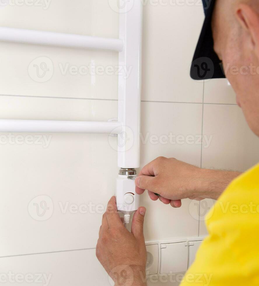 Repair in the bathroom. Male hands of a worker with a screwdriver. Plumber sets a heated towel rail. The interior of the walls with light tiles. photo