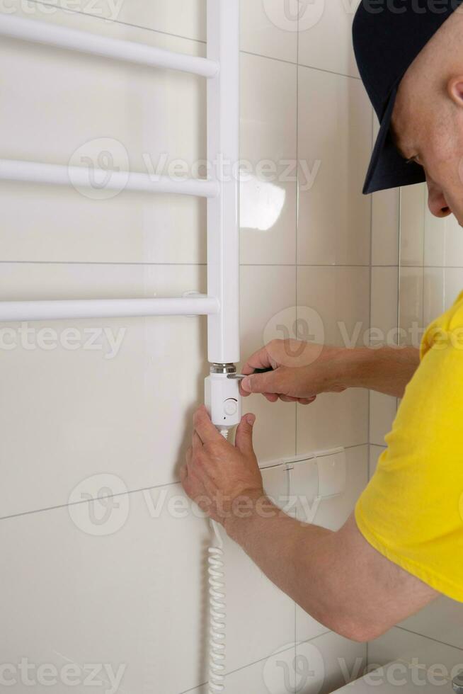 Repair in the bathroom. Male hands of a worker with a screwdriver. Plumber sets a heated towel rail. The interior of the walls with light tiles. photo