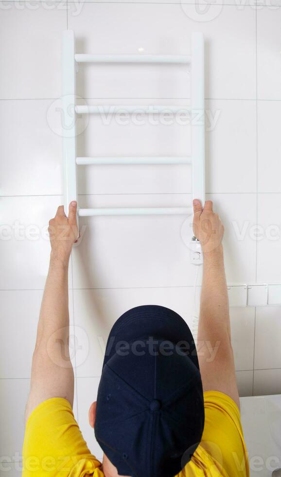 Renovation in the bathroom. A plumber in a cap and yellow shirt installs a heated towel rail. Interior walls with light tiles. High quality photo