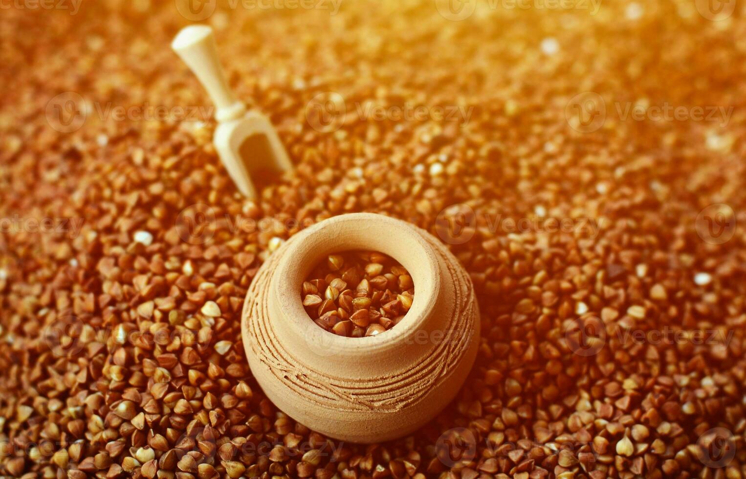Background image of a large pile of buckwheat, in the middle of which lies a small jug and a wooden spatula for cereals photo