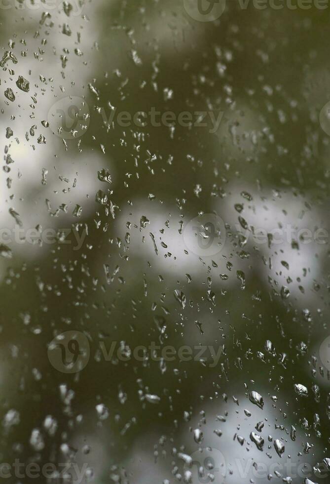 A photo of rain drops on the window glass with a blurred view of the blossoming green trees. Abstract image showing cloudy and rainy weather conditions