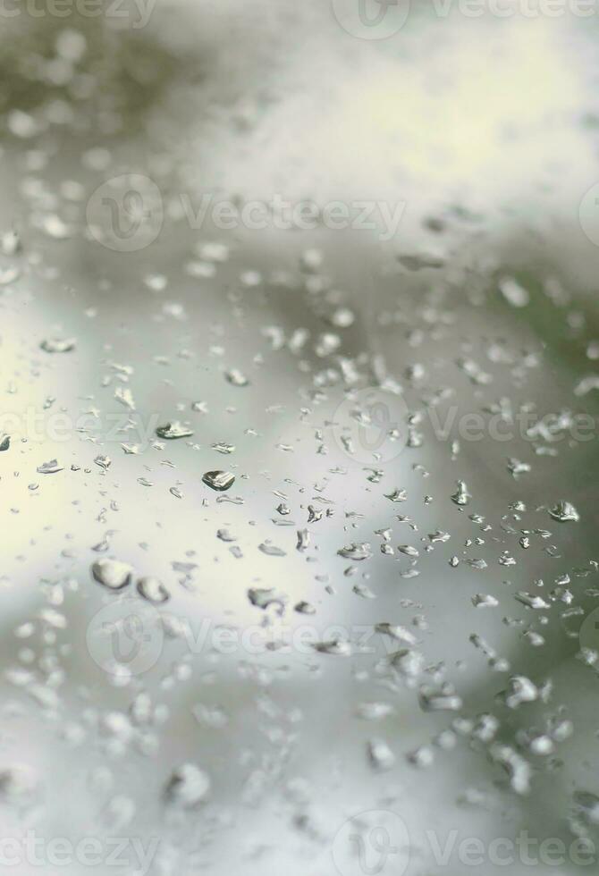 A photo of rain drops on the window glass with a blurred view of the blossoming green trees. Abstract image showing cloudy and rainy weather conditions