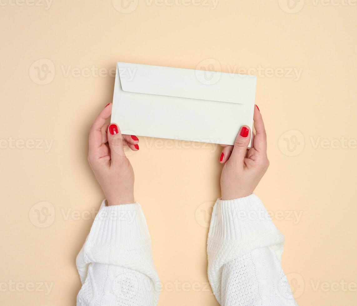 Two female hands holding a white rectangular envelope on a beige background, top view photo