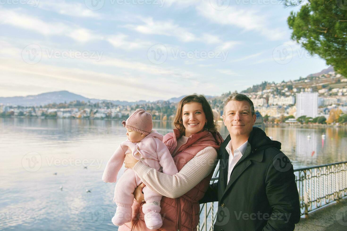 Outdoor portrait of happy young couple with adorable baby girl enjoying nice view of winter lake Geneva or Lac Leman, Montreux, Switzerland photo