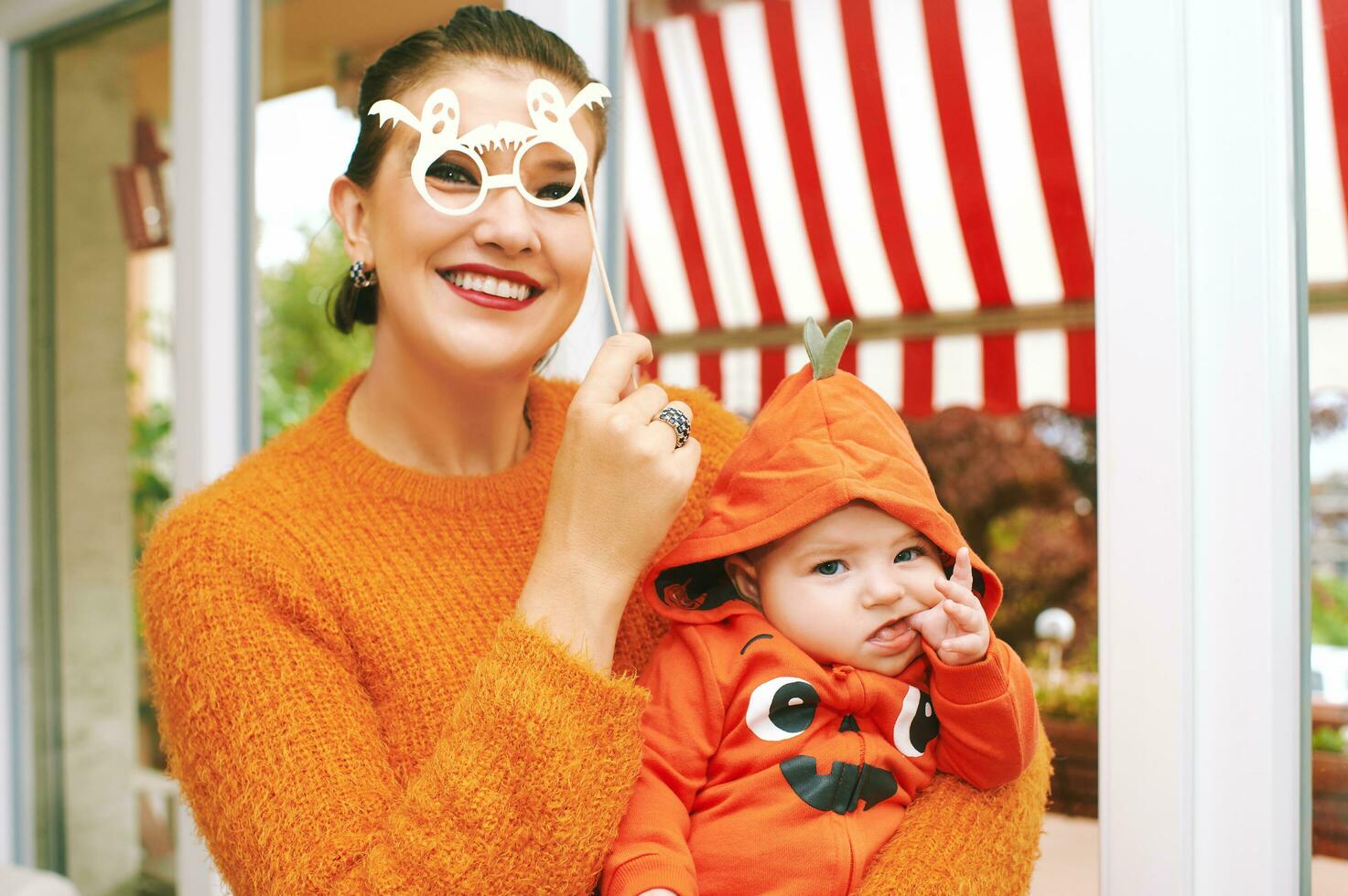 Halloween party with baby, mother holding child wearing pumpkin costume, posing with ghost paper glasses photo