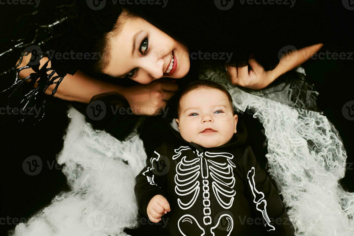 Halloween portrait of adorable baby wearing skeleton costume, posing on black background covered with spider web, mother wearing witch costume lying next to baby photo