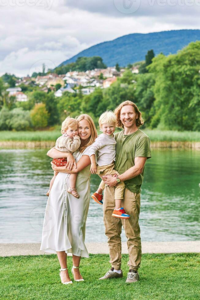 Outdoor portrait of beautiful family, young couple with preschooler boy and toddler girl posing next to lake or river photo