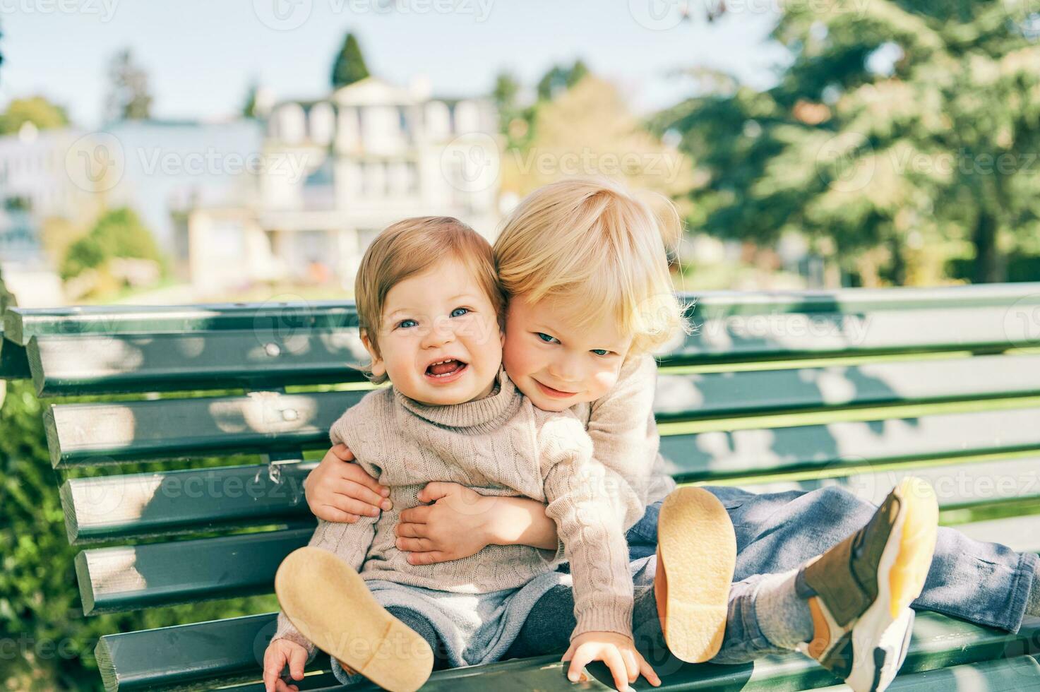 Outdoor portrait of two adorable siblings resting on the bench in sunny park, preschooler brother hugging sweet toddler sister photo