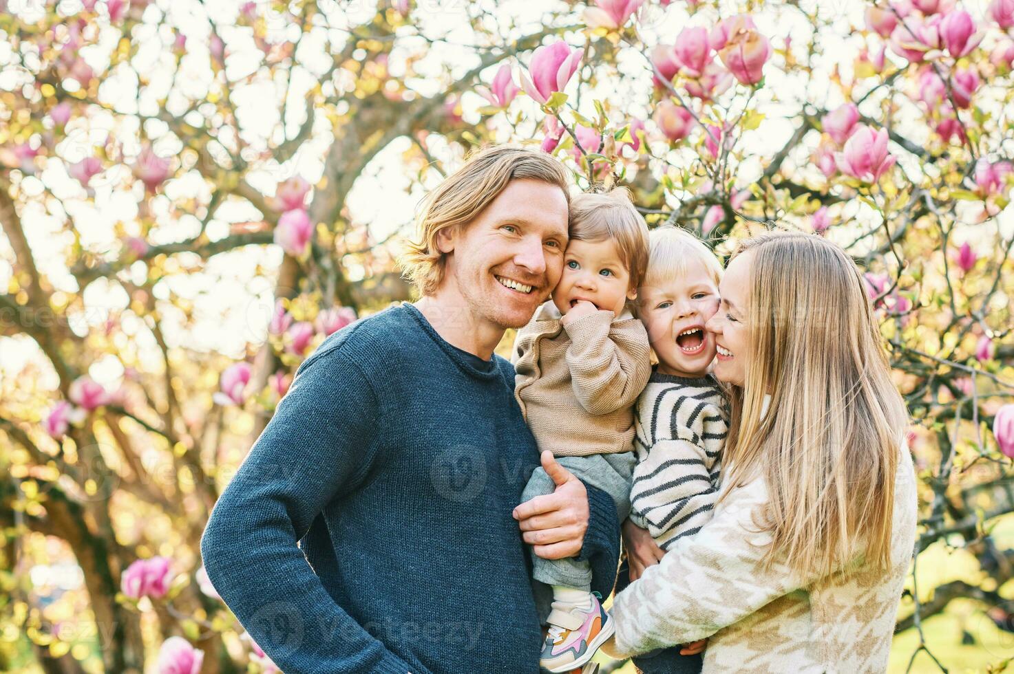 Outdoor portrait of happy young family playing in spring park under blooming magnolia tree, lovely couple with two little children having fun in sunny garden photo