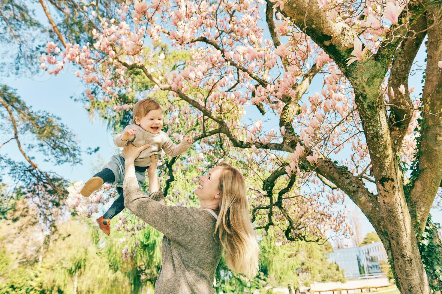 Outdoor portrait of happy young mother with adorable baby girl under blooming spring tree, having good time in sunny park photo