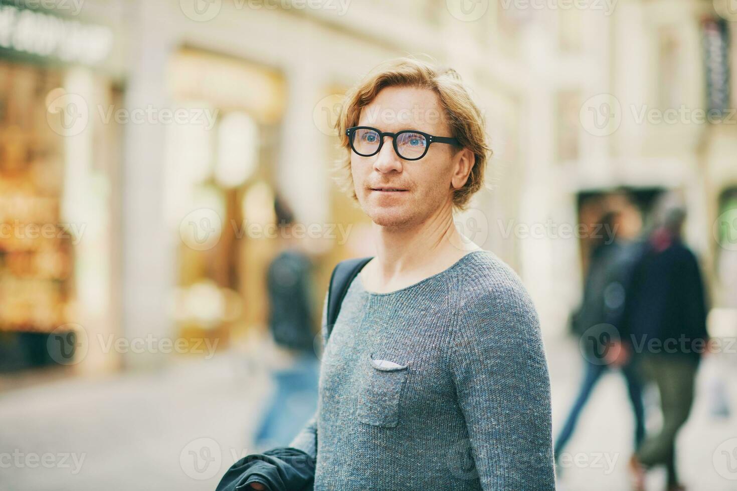Outdoor portrait of handsome red-haired man wearing eyeglasses posing on city street photo