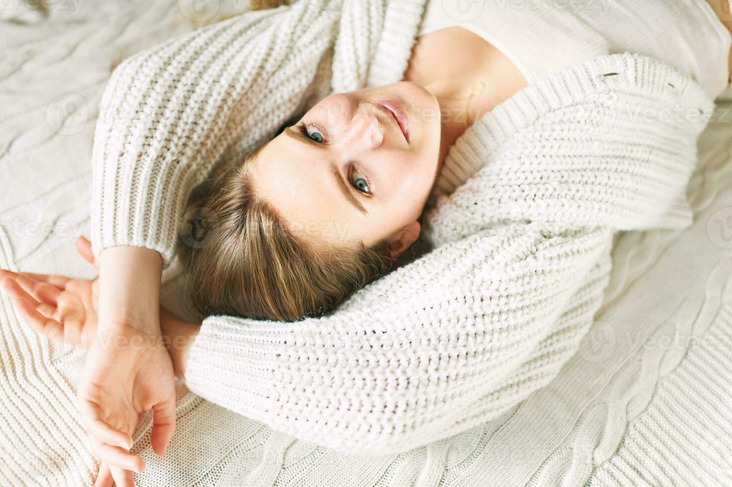 Portrait of young girl lying on white blanket, looking straight at camera, wearing warm knitted cardigan photo