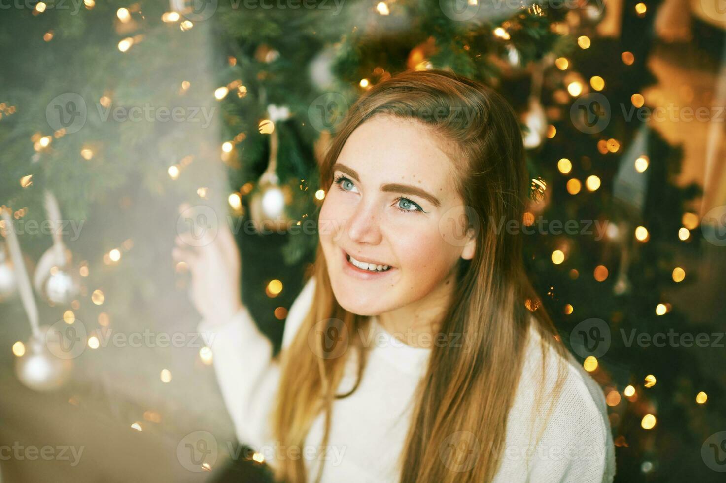 Young pretty girl looking up throught the window, christmas tree on background photo