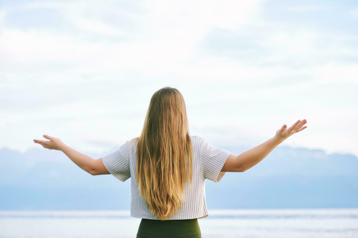 Outdoor portrait of young beautiful woman practicing yoga by the lakeYoung blond woman with arms wide open enjoy landscape of cloudy mountains, back view photo