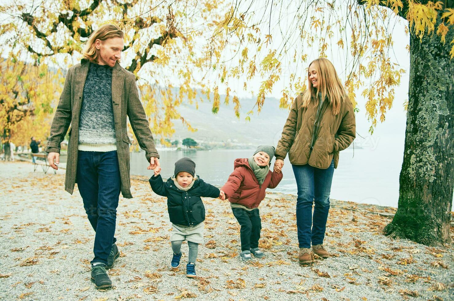 al aire libre retrato de joven contento familia de cuatro, madre y padre jugando con niños en otoño parque por el lago, frío clima foto