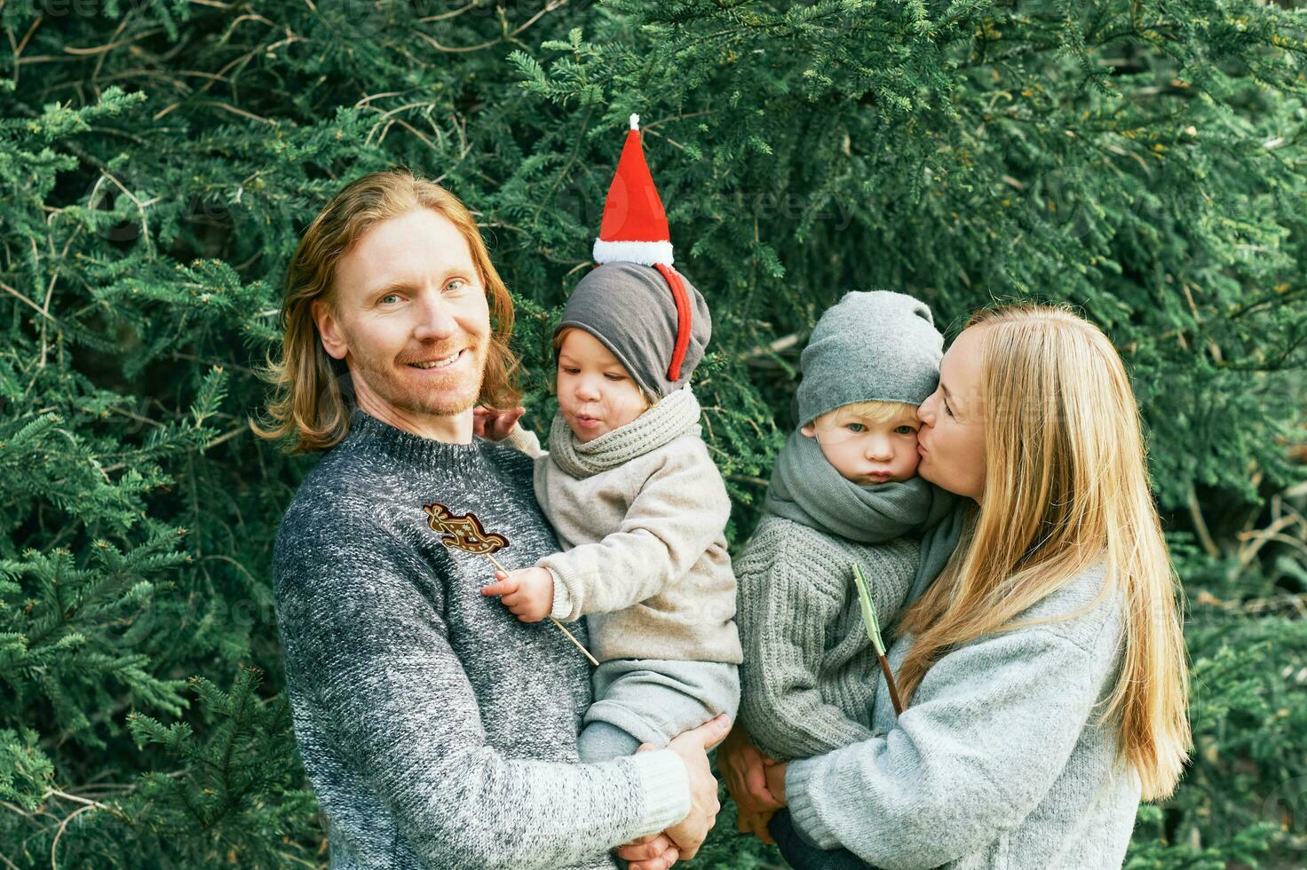 Outdoor portrait of beautiful happy young family of 4 posing in pine forest, wearing warm pullovers, couple with toddler boy and baby girl having good time at nature, cold weather, Christmas theme photo