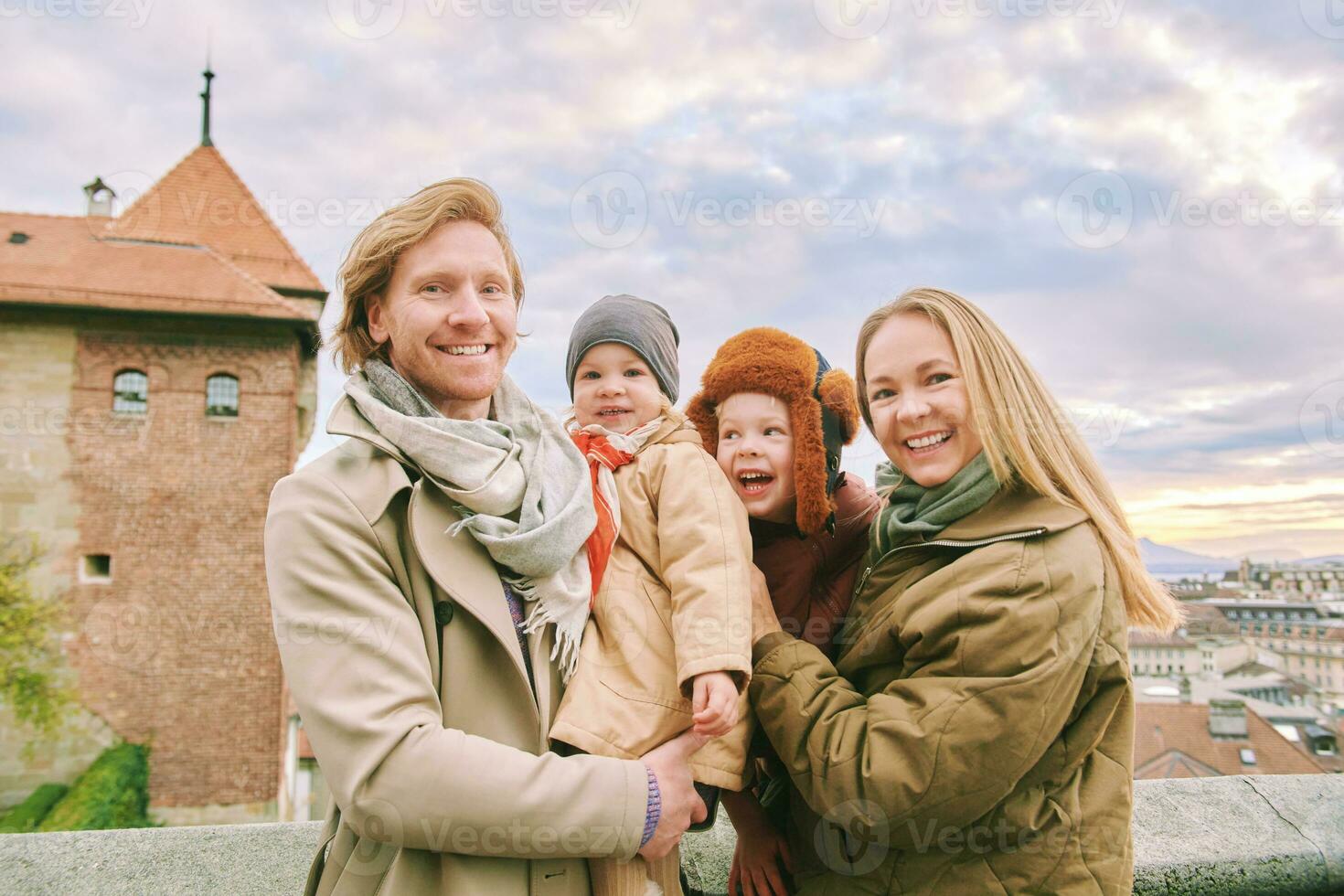 Outdoor portrait of happy family of four, young couple with two little children, cold weather, old european city on background photo