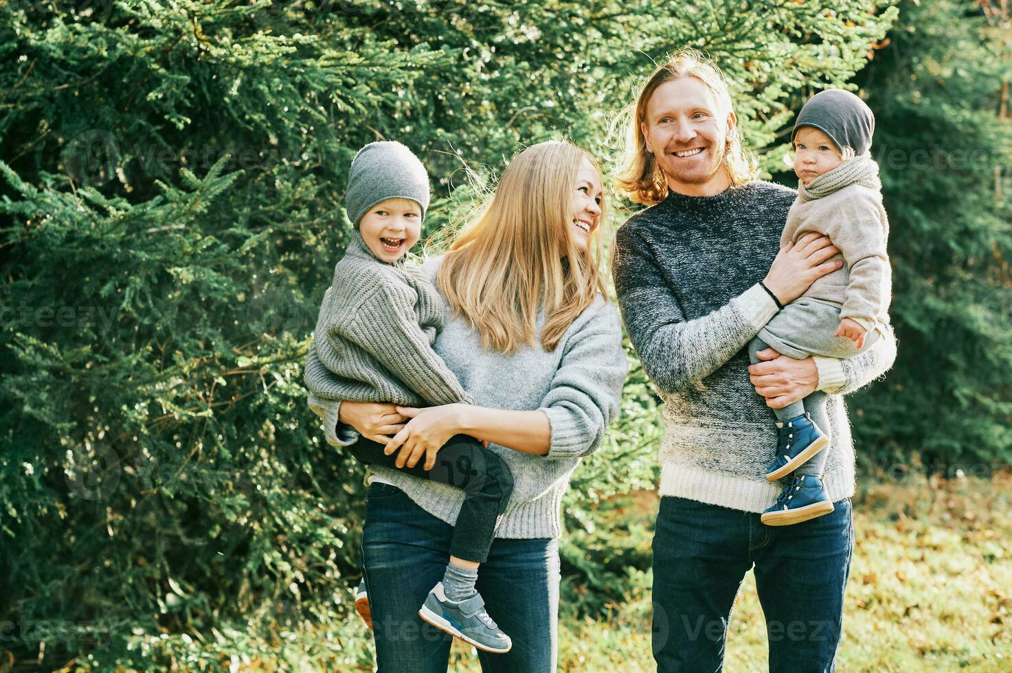 Outdoor portrait of beautiful happy young family of 4 posing in pine forest, wearing warm pullovers, couple with toddler boy and baby girl having good time at nature, cold weather photo