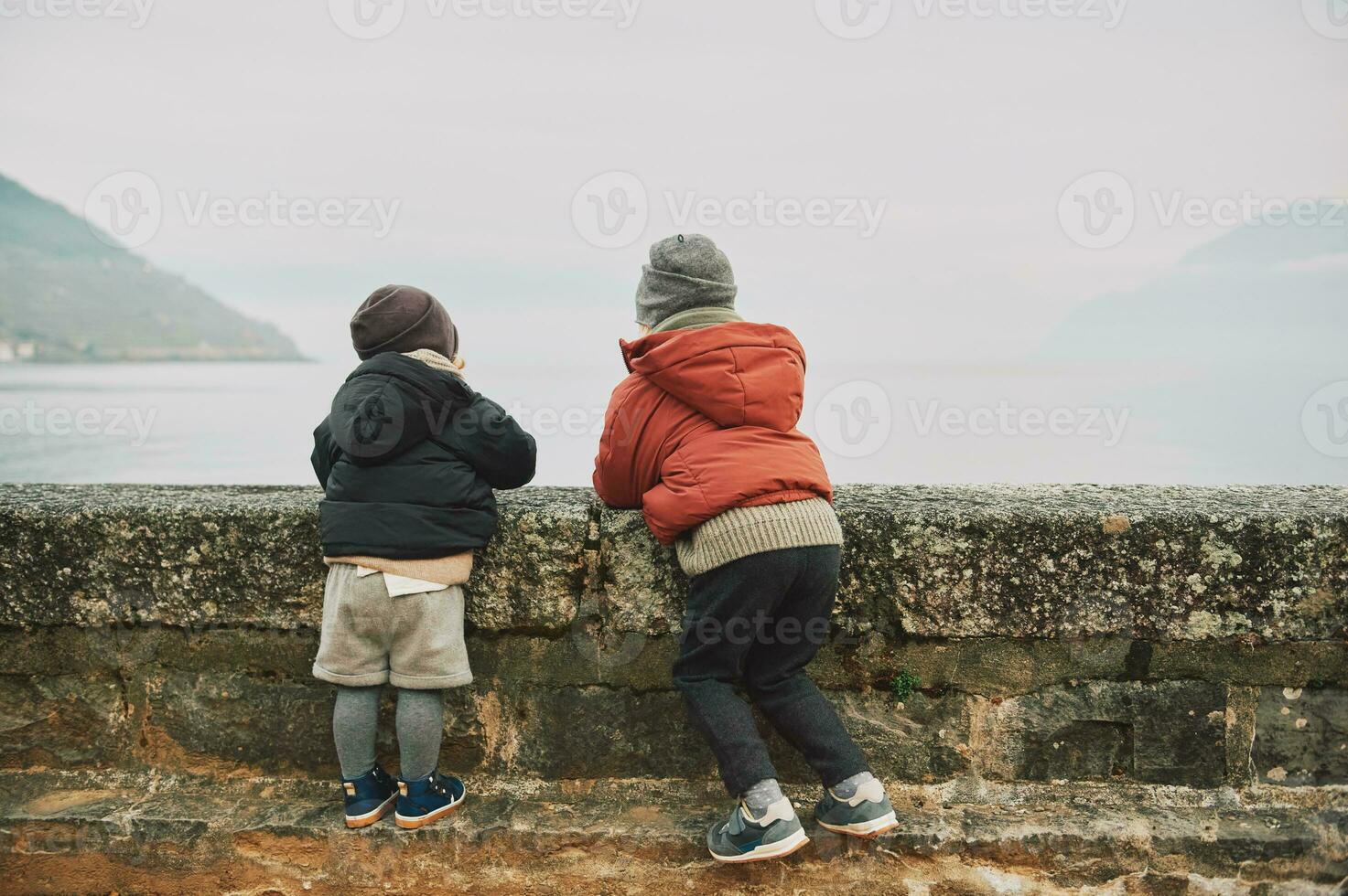 Outdoor portrait of two funny kids playing by the lake on a cold autumn day, wearing warm winter jackets, back view photo