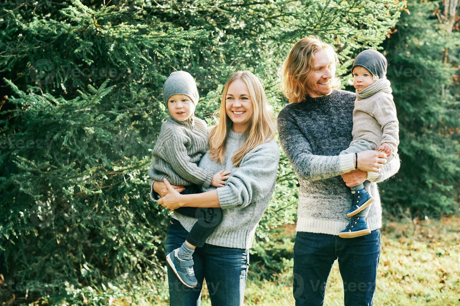 Outdoor portrait of beautiful happy young family of 4 posing in pine forest, wearing warm pullovers, couple with toddler boy and baby girl having good time at nature, cold weather photo