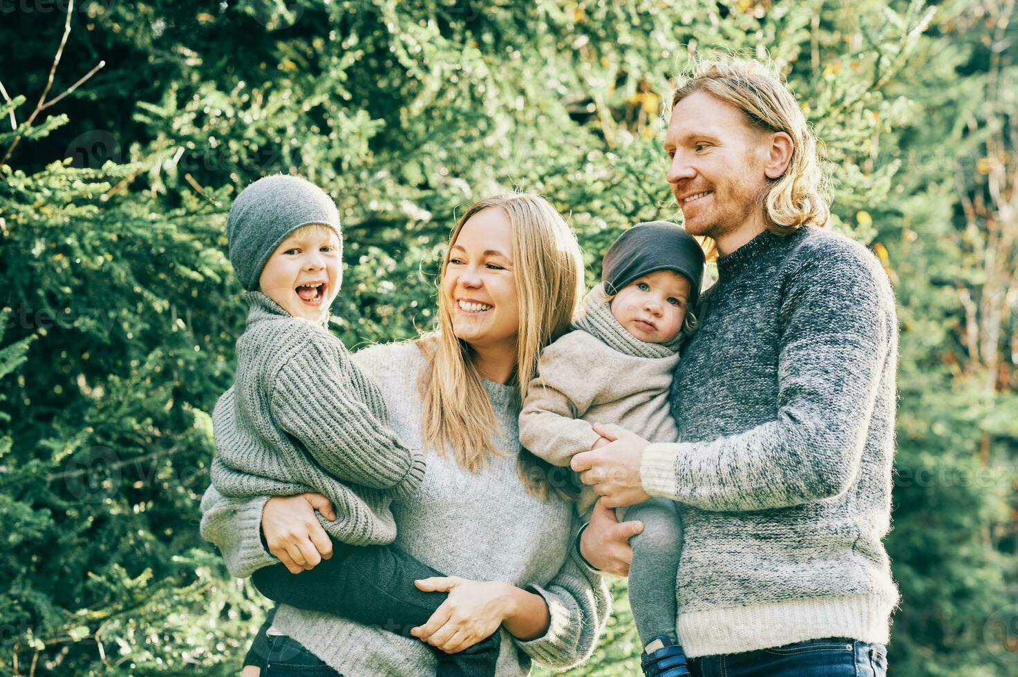 Outdoor portrait of beautiful happy young family of 4 posing in pine forest, wearing warm pullovers, couple with toddler boy and baby girl having good time at nature, cold weather photo