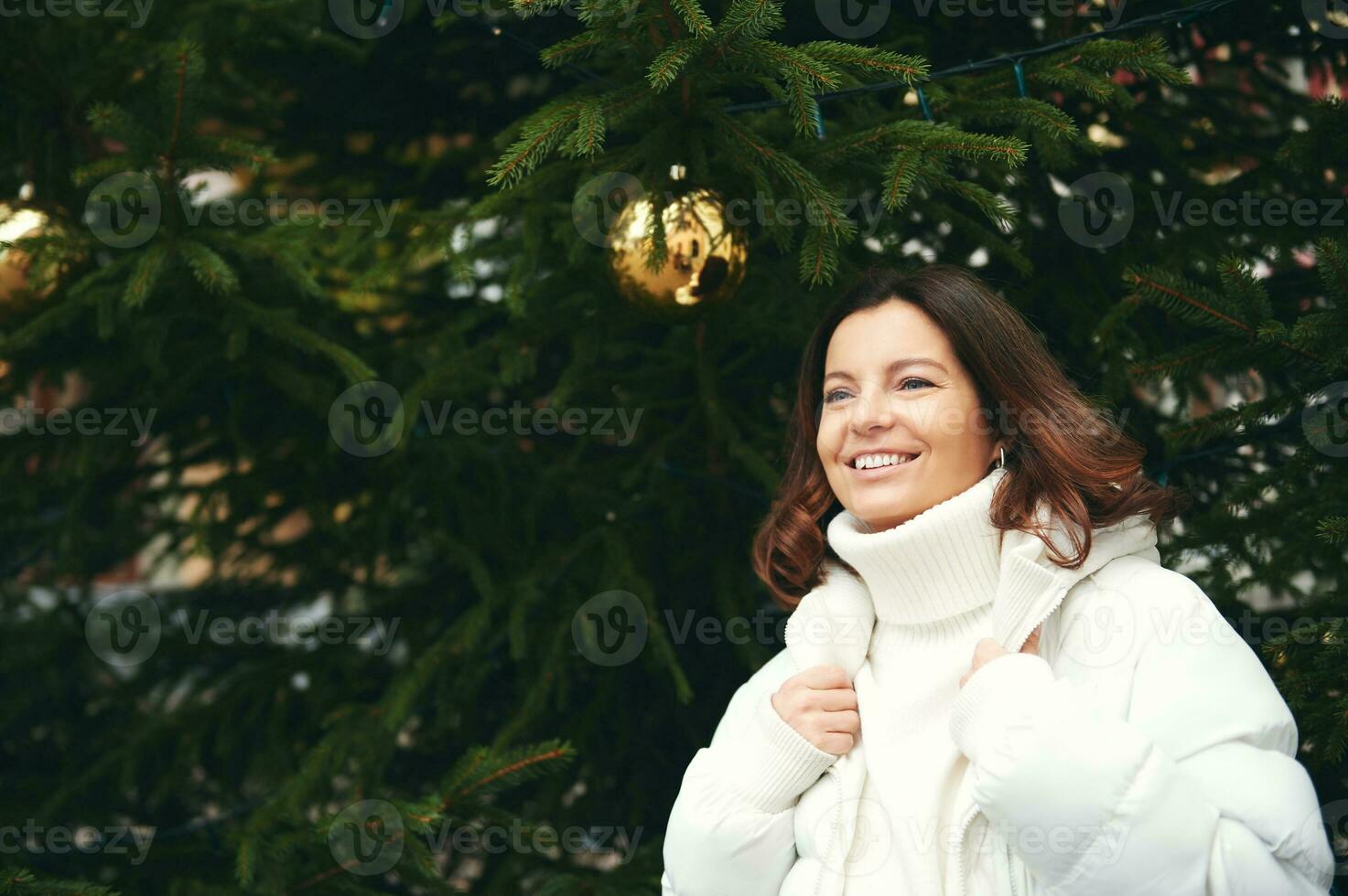 Outdoor portrait of happy woman next to green Christmas tree, wearing white pullover and jacket photo