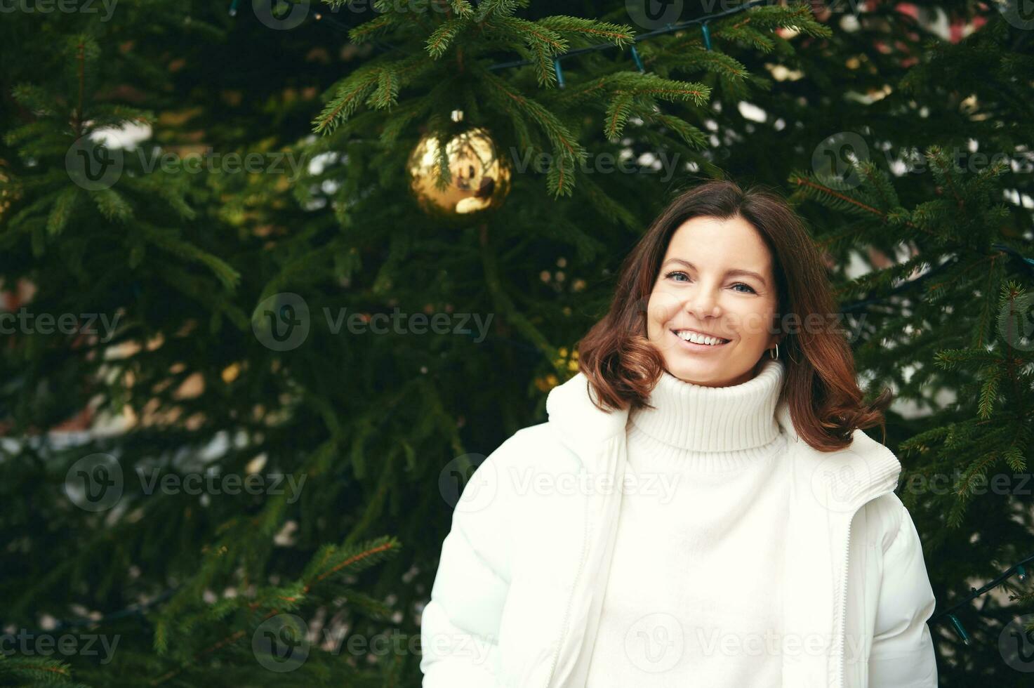 Outdoor portrait of happy woman next to green Christmas tree, wearing white pullover and jacket photo