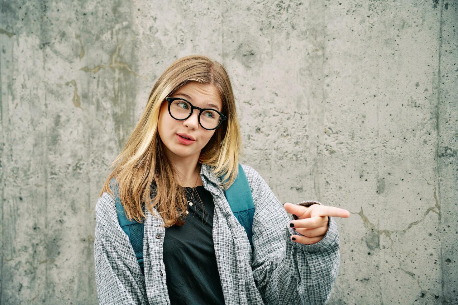 Outdoor portrait of young teenage kid girl wearing glasses and backpack, posing on grey wall background, talking and pointing finger to the side photo