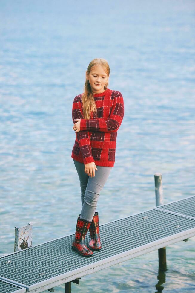 Outdoor portrait of cute little girl posing next to lake on dock, wearing red sweater and rain boots, cold fall or spring weather photo