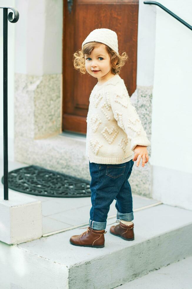 Outdoor portrait of adorable toddler girl wearing white knitted pullover and hat, dark blue denim jeans and brown retro shoes, fashion for little children photo