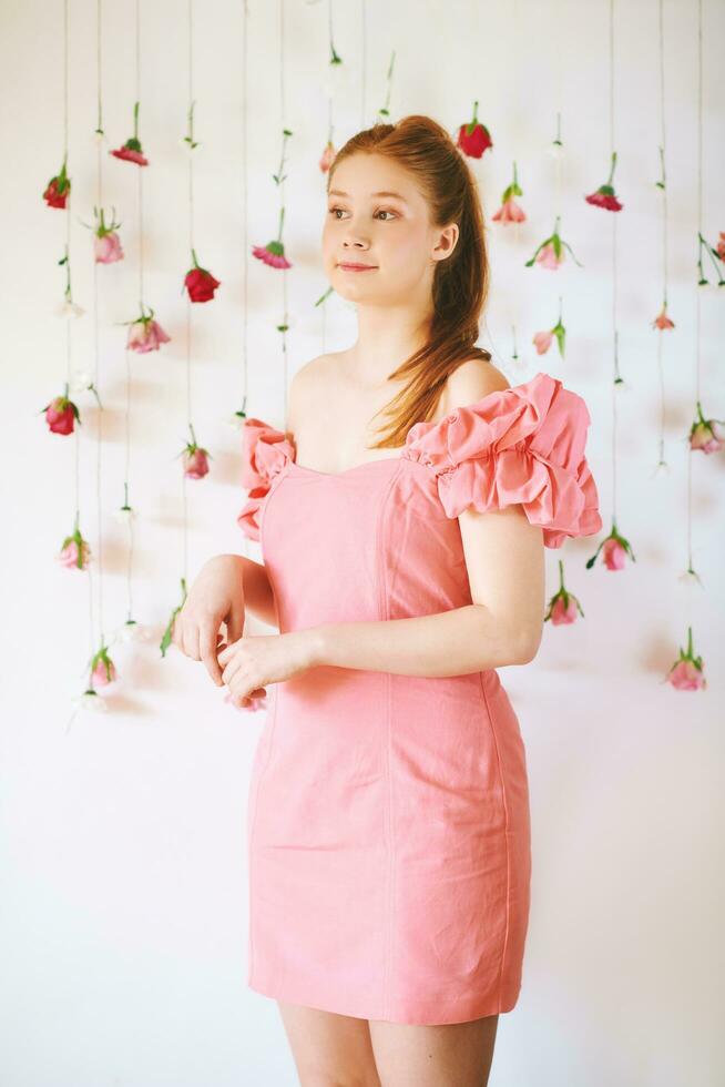 Studio portrait of pretty young teenage 15 - 16 year old red-haired girl wearing pink coral dress, posing on white background with hanging flowers, beauty and fashion concept photo