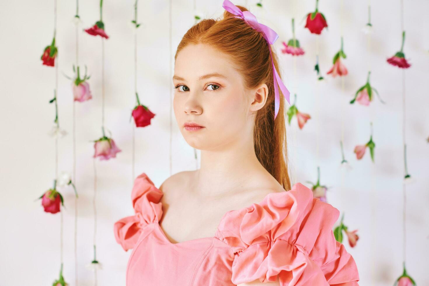 Studio portrait of pretty young teenage 15 - 16 year old red-haired girl wearing pink coral dress, posing on white background with hanging flowers, beauty and fashion concept photo