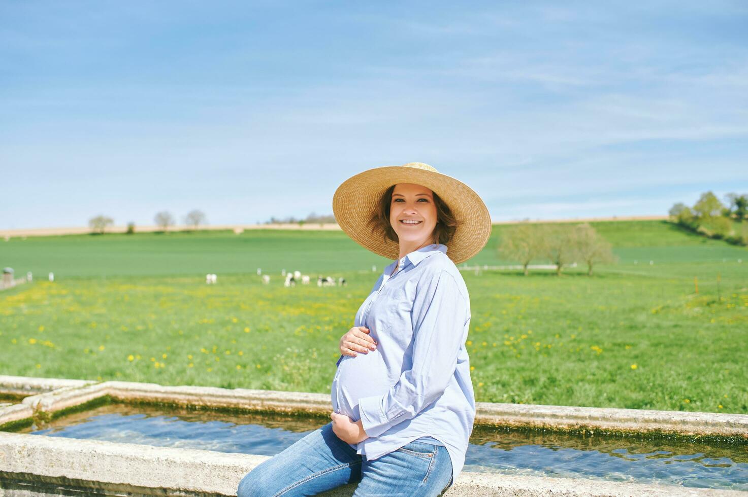 Outdoor portrait of happy young pregnant woman enjoying nice day in countryside, sitting on watering fountain, green pasture with cows on background photo