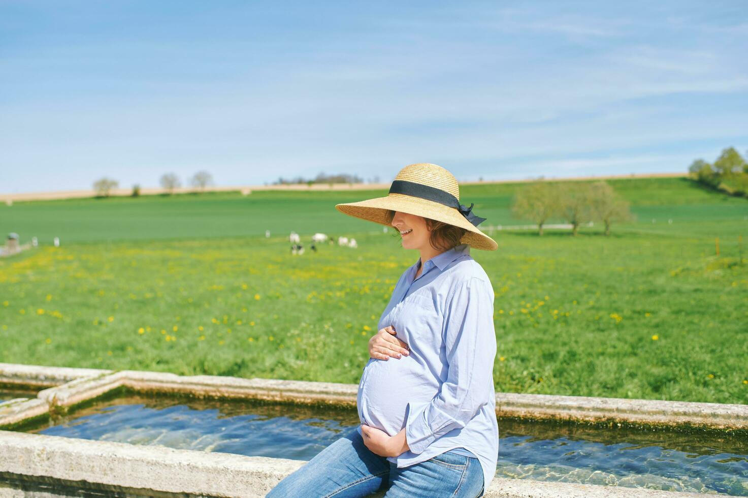 Outdoor portrait of happy young pregnant woman enjoying nice day in countryside, sitting on watering fountain, green pasture with cows on background photo