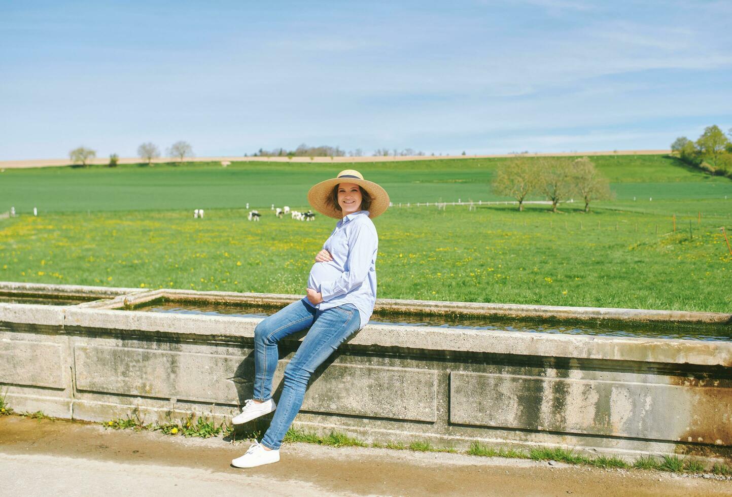Outdoor portrait of happy young pregnant woman enjoying nice day in countryside, sitting on watering fountain, green pasture with cows on background photo