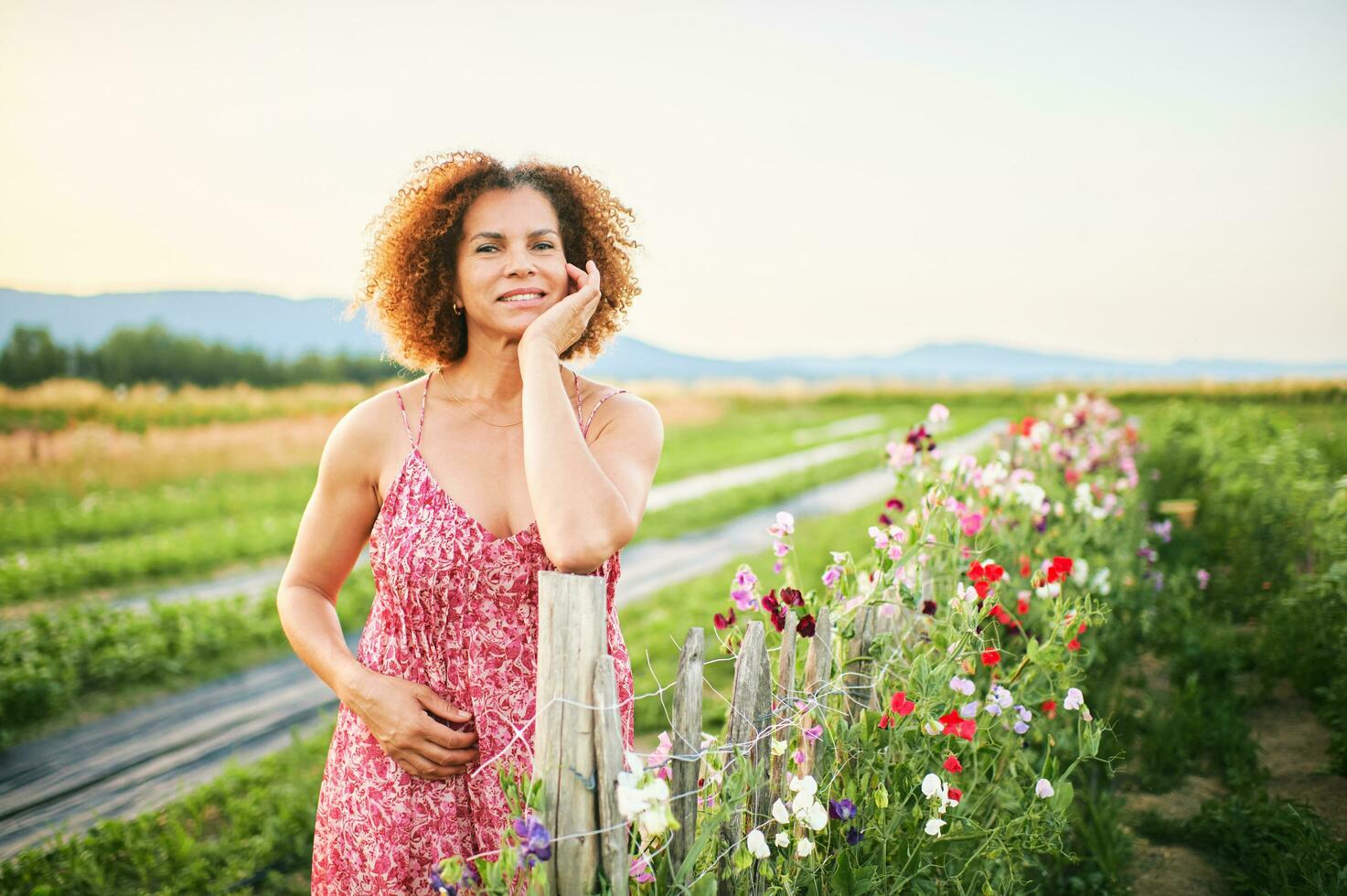 Outdoor portrait of beautiful middle age woman enjoying nice suuny evening in countryside, leaning on flower fence, wearing red dress photo