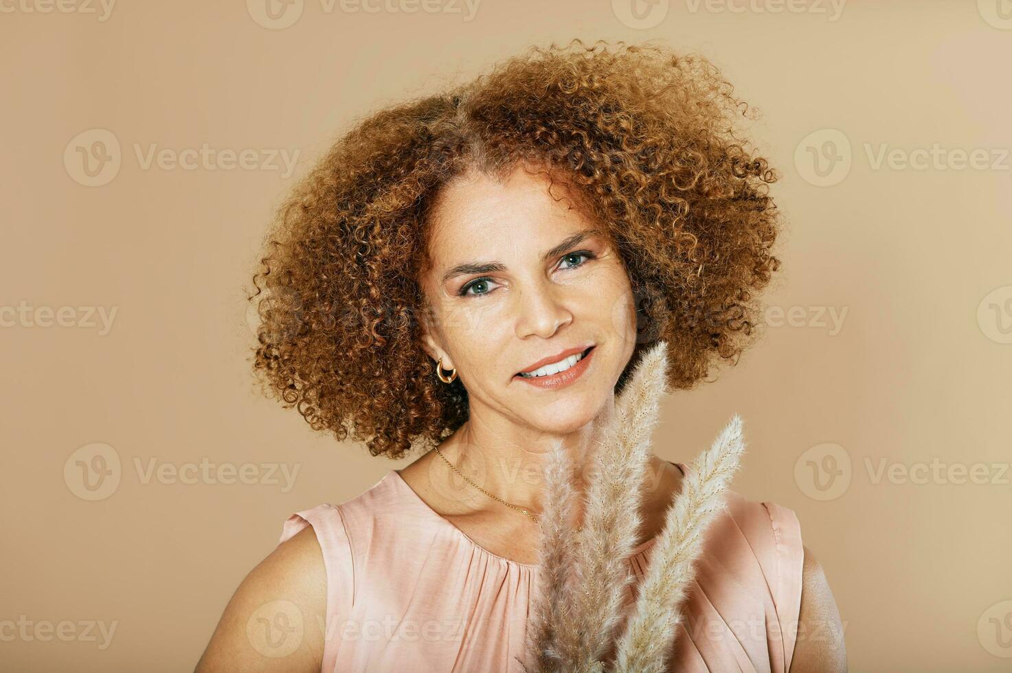 Fashion studio portrait of stylish middle age woman posing on beige background, 50 - 55 year old lady with curly hair holding pampas grass next to face photo