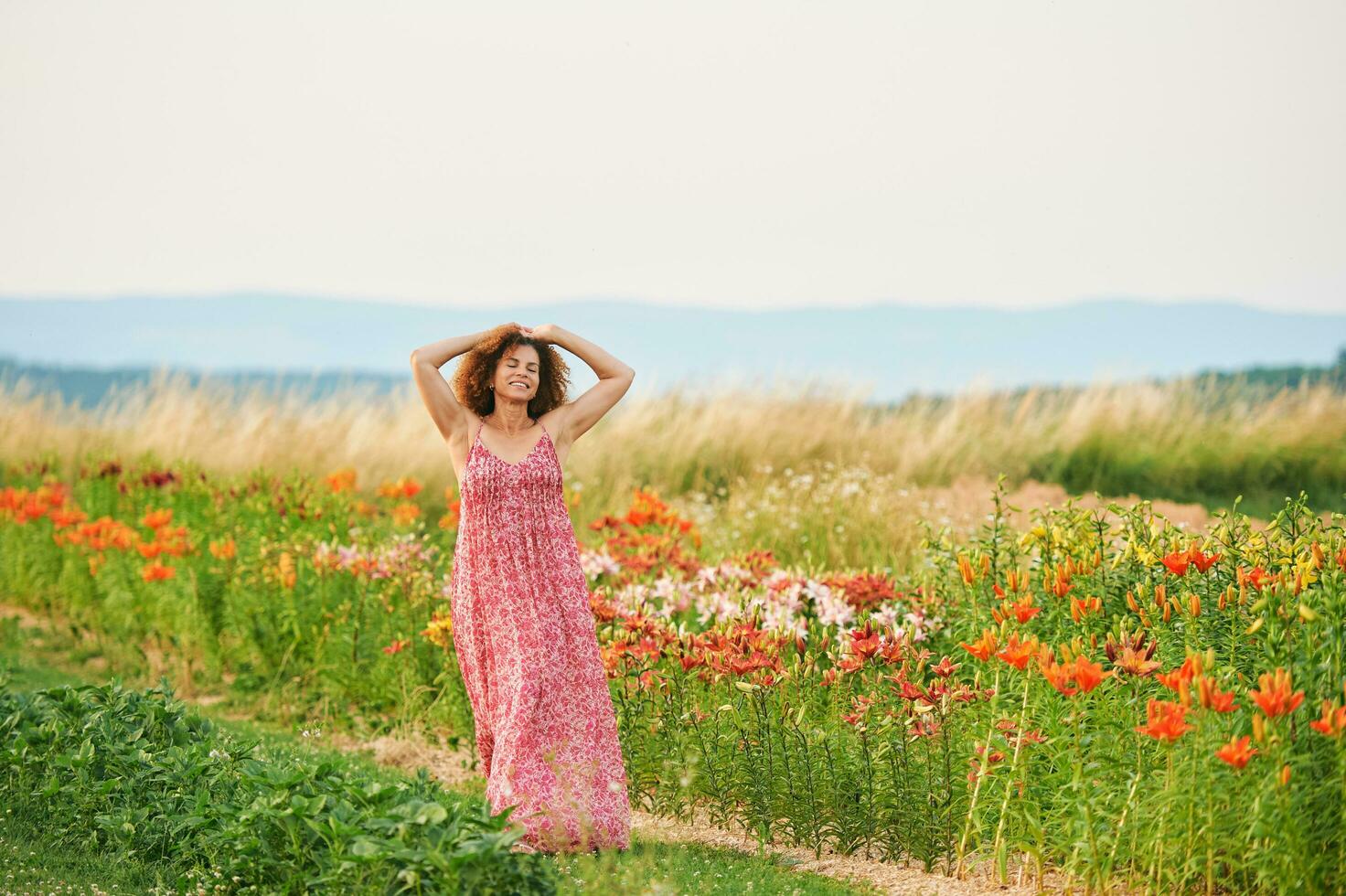 Outdoor portrait of beautiful middle age woman enjoying nice suuny evening in countryside, leaning on flower fence, wearing red dress photo