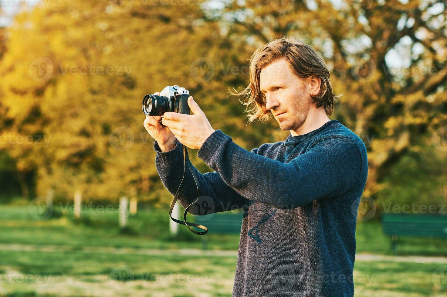 Outdoor portrait of handsome young man taking pictures on green sunny park with old vintage film camera photo