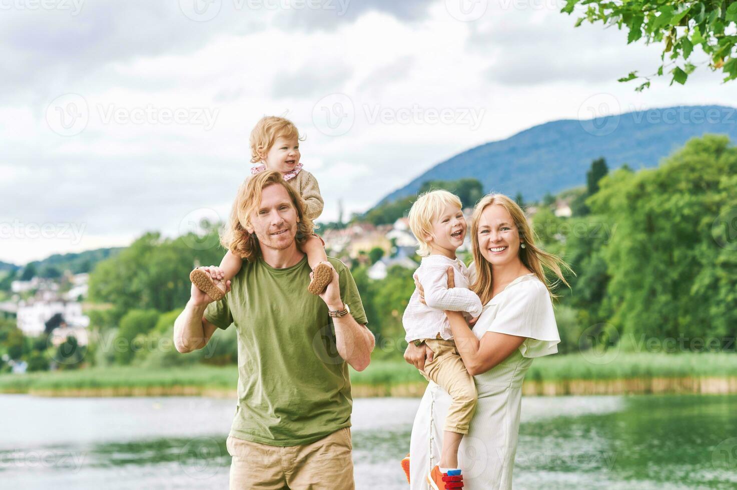 Outdoor portrait of beautiful family, young couple with preschooler boy and toddler girl posing next to lake or river photo