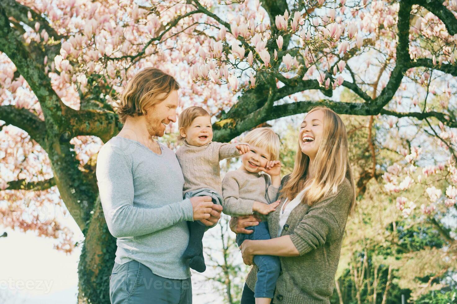 al aire libre retrato de contento joven familia jugando en primavera parque debajo floreciente magnolia árbol, encantador Pareja con dos pequeño niños teniendo divertido en soleado jardín foto