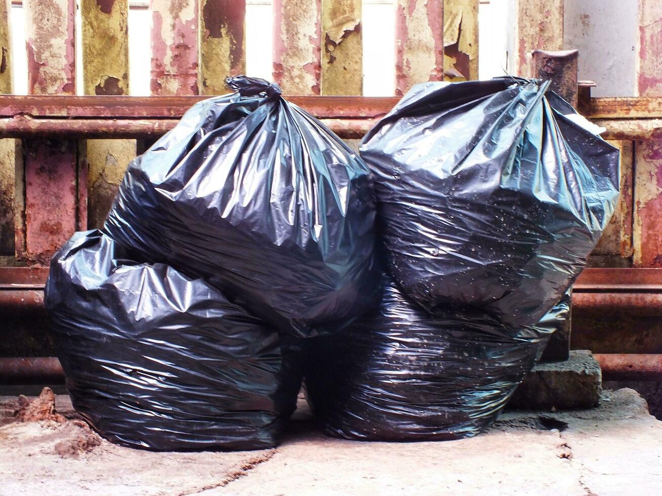 Black plastic bags of rubbish pile beside an old rusty french. Pile of black garbage on the footpath at side road in big city, pollution trash photo