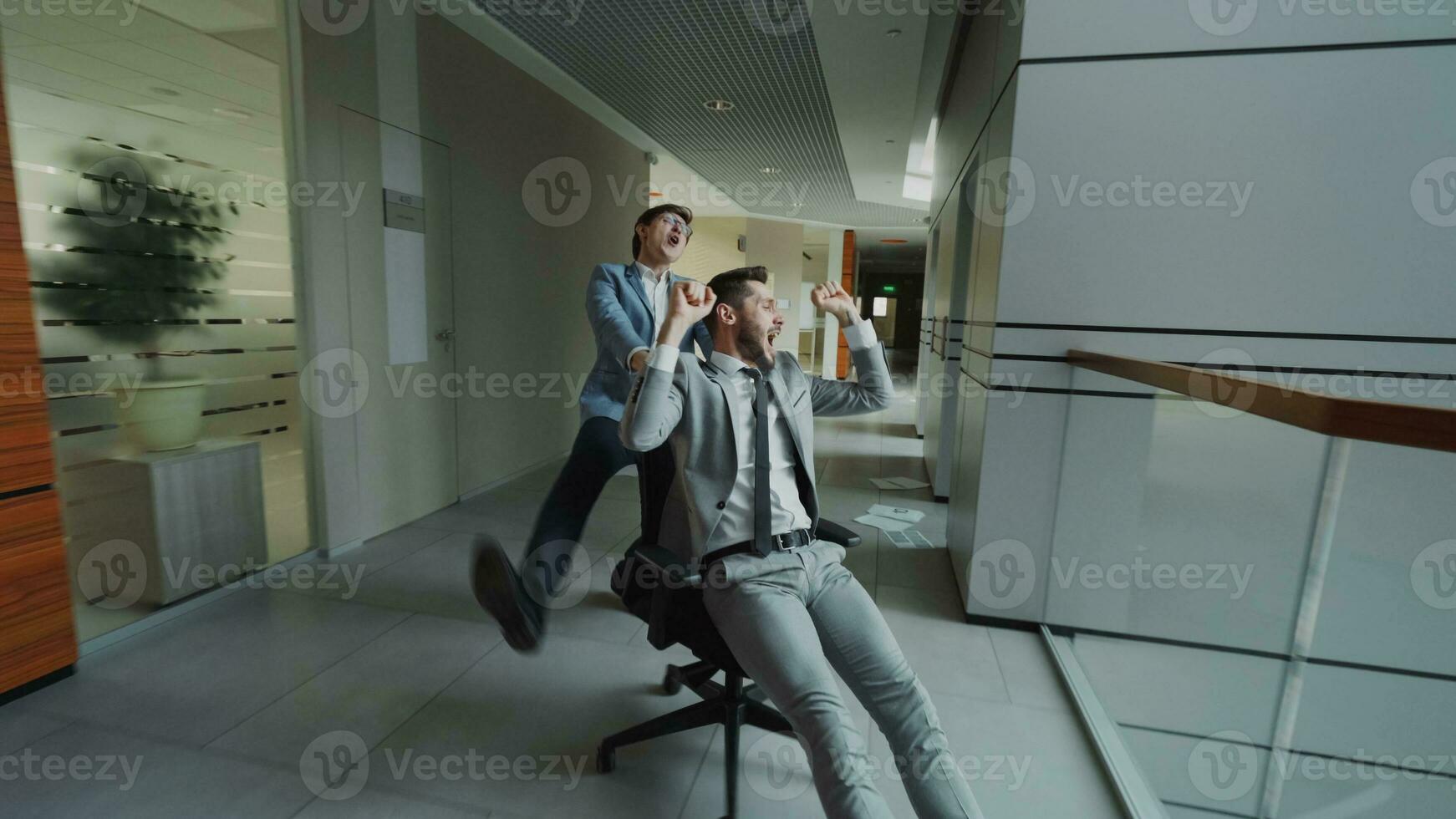 Two crazy businessmen riding office chair and throwing papers up while having fun in lobby of modern business center indoors photo