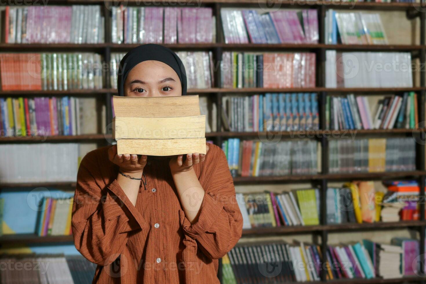 Portrait of Asian hijab woman covering her head with a pile of books in front of library bookshelf. Muslim girl reading a book. Concept of literacy and knowledge photo