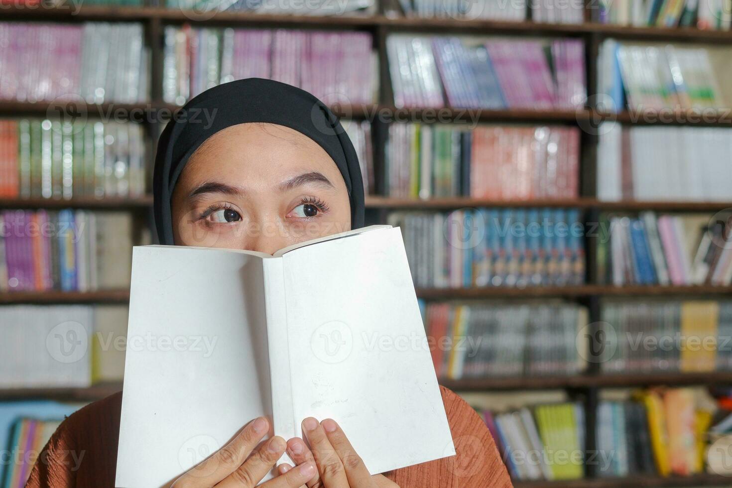 Portrait of Asian hijab woman covering her half head with a book in front of library bookshelf. Muslim girl reading a book. Concept of literacy and knowledge photo